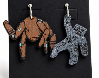 May include: A pair of mismatched dangle earrings featuring a copper-colored robotic figure with blue accents on the left and a textured, dark gray mechanical-patterned figure on the right, both suspended from silver-toned metal hoops.