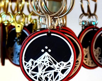 May include: Keychains with a black circular design featuring a white mountain range graphic, a red border, and a silver keyring. The keychains are hanging from a display, with several in focus.