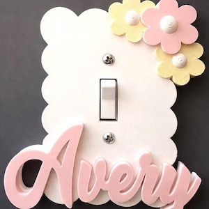 May include: A white scalloped light switch plate featuring the name Avery in raised pink script lettering at the bottom. The top right corner is decorated with three dimensional pastel yellow and pink flowers with white centers.