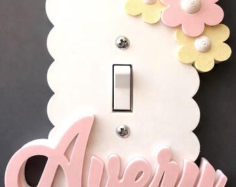 May include: A white scalloped light switch plate featuring the name Avery in raised pink script lettering. The plate is decorated with three dimensional pastel yellow and pink flowers in the top right corner and two silver metal screws.