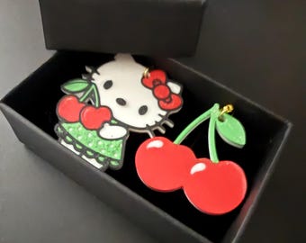 May include: Two acrylic pins in a black box. One pin is a white cat with a red bow and cherries. The other pin is a pair of red cherries with green stems and leaves. The pins are likely for a backpack or jacket.