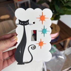 May include: A white scalloped light switch plate featuring a black cat silhouette on the left side and three starburst shapes on the right. The starbursts are arranged vertically, with two orange starbursts at the top and bottom and one teal starburst in the centre. The plate has a rectangular cutout in the middle and a small circular hole at the top.