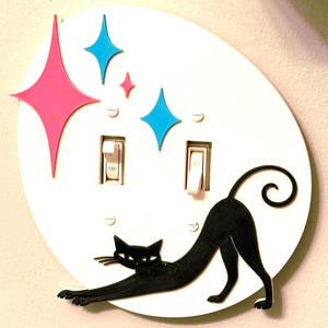 May include: A white circular double light switch plate featuring a black cat silhouette stretching across the bottom. The design includes one large pink starburst and two smaller blue starburst accents positioned above the two white toggle switches.