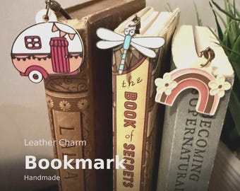 May include: Leather charm bookmarks in the image include a camper, dragonfly, and rainbow design. The camper is pink and white with a red door. The dragonfly is white and blue. The rainbow is brown and pink.
