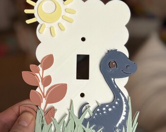 May include: A decorative light switch cover featuring a whimsical design. The cover includes a yellow sun, a blue dinosaur, and green grass. The background is white with a cloud-like shape. The switch opening is a standard size.