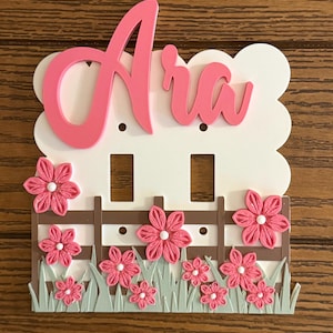 Op de afbeelding: Een witte afdekplaat voor een dubbele lichtschakelaar met de naam "Ara" in grote roze schrijfletters aan de bovenkant. Het onderste gedeelte toont een bruin hek met lichtgroen gras en diverse roze stoffen bloemen met witte harten.