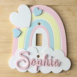 Op de afbeelding: Een regenboogvormige lichtschakelaarafdekking met pastelroze, gele en blauwe bogen. Het ontwerp bevat een groot wit hart, een kleiner blauw hart en een witte wolkenbasis met de naam Sophia in verhoogde roze cursieve letters.