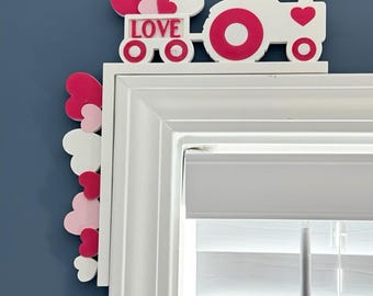 May include: A white wooden tractor with pink heart accents and the word "LOVE" on the side. The tractor is decorated with pink and white hearts, perfect for Valentine's Day or a love-themed decoration. The tractor is placed above a window.