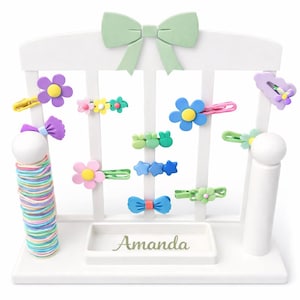 May include: A white wooden hair accessory organizer featuring a pastel green bow at the top and a tray with the name Amanda printed on it. The stand holds a stack of colorful elastic hair ties and various hair clips shaped like flowers and bows.