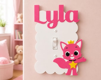 May include: A white scalloped light switch cover featuring the name Lyla in pink letters at the top and a pink cartoon fox character wearing a yellow crown and a yellow star on its chest attached to the bottom right corner of the plate.
