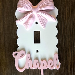 Op de afbeelding: Een witte rechthoekige lichtschakelaarafdekking met geschulpte randen, voorzien van een driedimensionale lichtroze strik aan de bovenkant en de naam Chapel in lichtroze cursief schrift aan de onderkant, met twee schroefgaten.