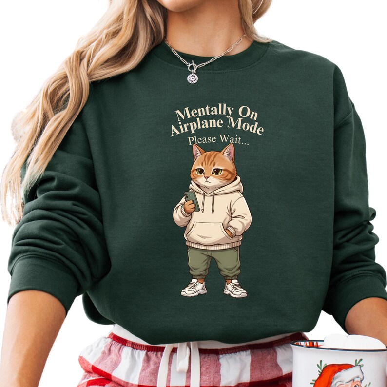 May include: Dark green sweatshirt with the text "Mentally On Airplane Mode Please Wait..." above a cartoon cat wearing a hoodie, pants, and sneakers, holding a phone. The cat is orange and white.
