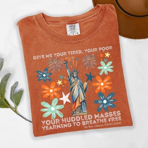 Puede incluir: Camiseta de color naranja quemado con un gr&aacute;fico de la Estatua de la Libertad, fuegos artificiales y dise&ntilde;os florales. El texto "GIVE ME YOUR TIRED, YOUR POOR" y "YOUR HUDDLED MASSES YEARNING TO BREATHE FREE" est&aacute; impreso en la camiseta.