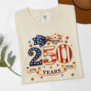 May include: Cream-colored t-shirt with a patriotic design. Features a bow, the number "250" in red, white, and blue, and the text "1776 YEARS 2026" on a banner. The shirt has the Comfort Colors brand label.
