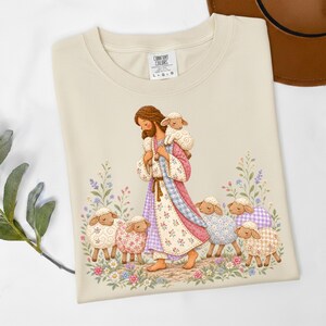 May include: A beige t-shirt featuring a graphic illustration of a figure with long brown hair and a beard, wearing a patterned robe and carrying a small lamb. The figure is surrounded by several sheep with floral patterns and colorful wildflowers.
