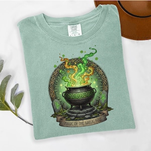 May include: A light green t-shirt featuring a cauldron design with green and gold smoke, surrounded by Celtic knotwork and runes. The text "MAGIC OF THE GAELIC SOUL" is displayed on a banner below the cauldron. The shirt has a crew neck and short sleeves.