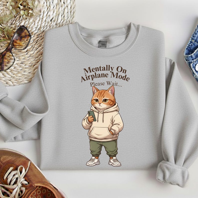 May include: A light gray sweatshirt featuring a cartoon cat wearing a hoodie and holding a phone. The text "Mentally On Airplane Mode Please Wait..." is printed above the cat. The cat is orange and white, wearing green pants and white sneakers.