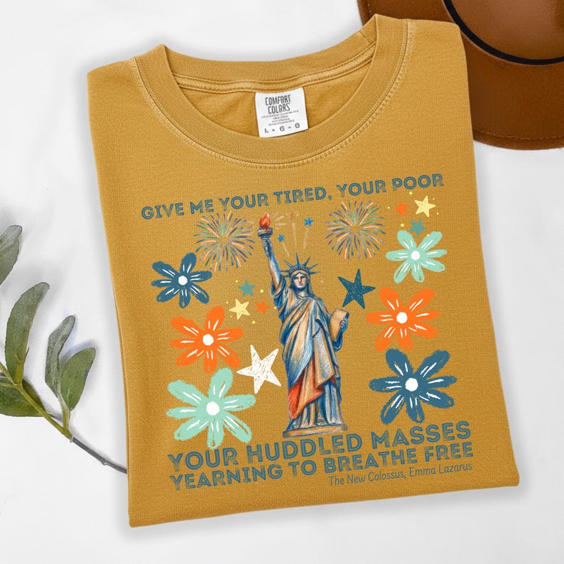 Puede incluir: Camiseta de color amarillo mostaza con un gr&aacute;fico de la Estatua de la Libertad, fuegos artificiales y flores en azul, naranja y blanco. El texto dice "GIVE ME YOUR TIRED, YOUR POOR" y "YOUR HUDDLED MASSES YEARNING TO BREATHE FREE."