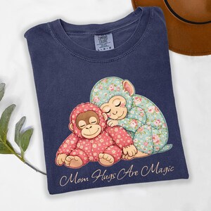 May include: Navy blue t-shirt with a graphic of two monkeys hugging, one in pink floral fabric and the other in blue floral fabric. The text "Mom Hugs Are Magic" is printed below the monkeys. The shirt is from Comfort Colors.