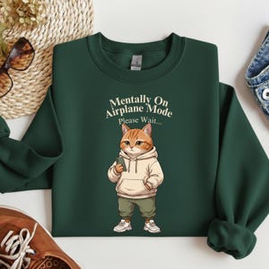 May include: Dark green sweatshirt with the text "Mentally On Airplane Mode Please Wait..." above a cartoon cat wearing a hoodie and holding a phone. The cat is orange and white, wearing green pants and white sneakers.
