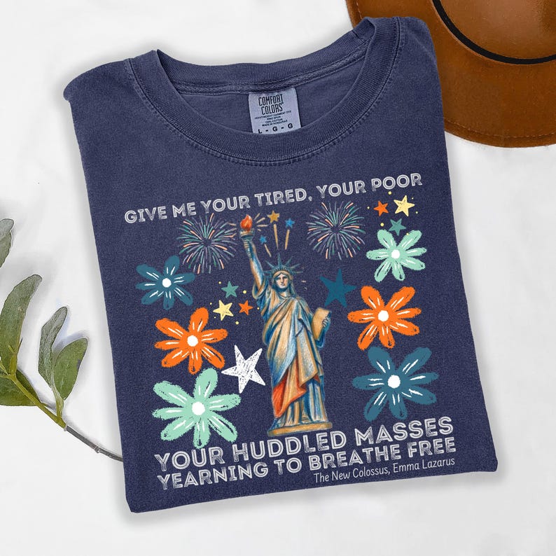Puede incluir: Camiseta azul marino con un dise&ntilde;o de la Estatua de la Libertad, rodeada de flores y fuegos artificiales. El texto "GIVE ME YOUR TIRED, YOUR POOR" y "YOUR HUDDLED MASSES YEARNING TO BREATHE FREE" est&aacute; impreso.