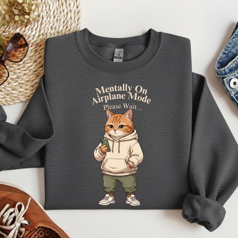 May include: Dark gray sweatshirt featuring a cartoon cat wearing a hoodie and holding a phone. The text "Mentally On Airplane Mode Please Wait..." is printed above the cat. The cat is wearing green pants and white sneakers.