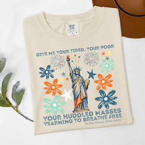 Puede incluir: Camiseta color crema con un gr&aacute;fico de la Estatua de la Libertad y el texto "GIVE ME YOUR TIRED, YOUR POOR" y "YOUR HUDDLED MASSES YEARNING TO BREATHE FREE". El dise&ntilde;o incluye flores y fuegos artificiales coloridos.