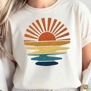 May include: Cream-colored t-shirt featuring a sunset design. The graphic depicts a sun with orange rays above horizontal bands of orange, yellow, teal, and navy blue, resembling a sunset over water. The shirt has short sleeves.