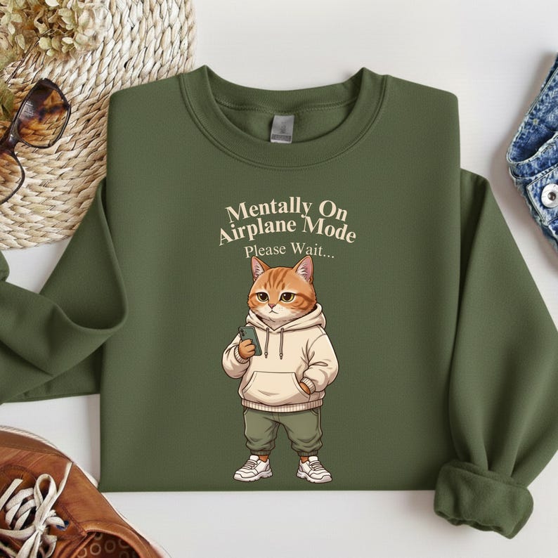 May include: Olive green sweatshirt with the text "Mentally On Airplane Mode Please Wait..." and a cartoon cat wearing a hoodie and holding a phone. The cat is orange and white, wearing a beige hoodie, green pants, and white sneakers.