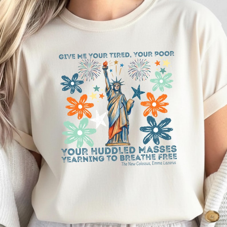 Puede incluir: Camiseta color crema con un dise&ntilde;o gr&aacute;fico de la Estatua de la Libertad, fuegos artificiales y flores. El texto incluye versos del poema de Emma Lazarus: "GIVE ME YOUR TIRED, YOUR POOR... YEARNING TO BREATHE FREE".