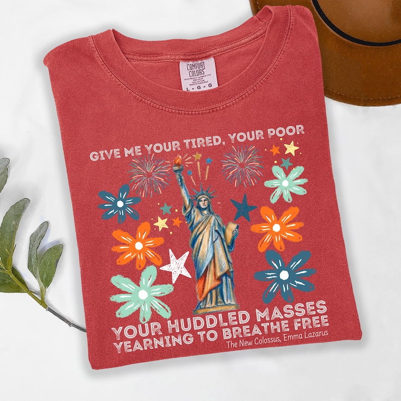Puede incluir: Una camiseta roja con un gr&aacute;fico de la Estatua de la Libertad, fuegos artificiales y flores. El texto dice: "GIVE ME YOUR TIRED, YOUR POOR, YOUR HUDDLED MASSES YEARNING TO BREATHE FREE." La camiseta es de un material suave.