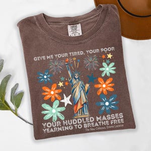 Puede incluir: Camiseta marr&oacute;n con un dise&ntilde;o de la Estatua de la Libertad, rodeada de flores y fuegos artificiales coloridos. El texto "GIVE ME YOUR TIRED, YOUR POOR" y "YOUR HUDDLED MASSES YEARNING TO BREATHE FREE" est&aacute; impreso en la camiseta.