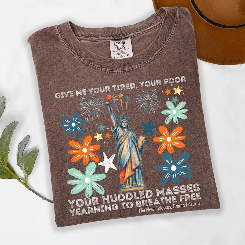 Puede incluir: Camiseta marr&oacute;n con un dise&ntilde;o de la Estatua de la Libertad, rodeada de flores y fuegos artificiales coloridos. El texto "GIVE ME YOUR TIRED, YOUR POOR" y "YOUR HUDDLED MASSES YEARNING TO BREATHE FREE" est&aacute; impreso en la camiseta.