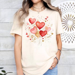May include: Cream-colored t-shirt featuring a watercolor design of heart-shaped balloons in shades of red, pink, and gold. The balloons are clustered together with small heart accents and gold foliage, creating a festive and romantic theme.