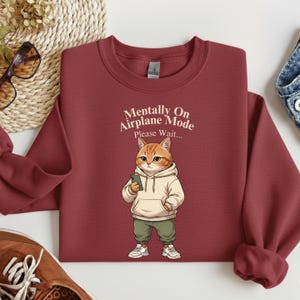 May include: A maroon sweatshirt with the text "Mentally On Airplane Mode Please Wait..." above a cartoon cat wearing a hoodie, green pants, and sneakers, holding a phone. The sweatshirt is folded.