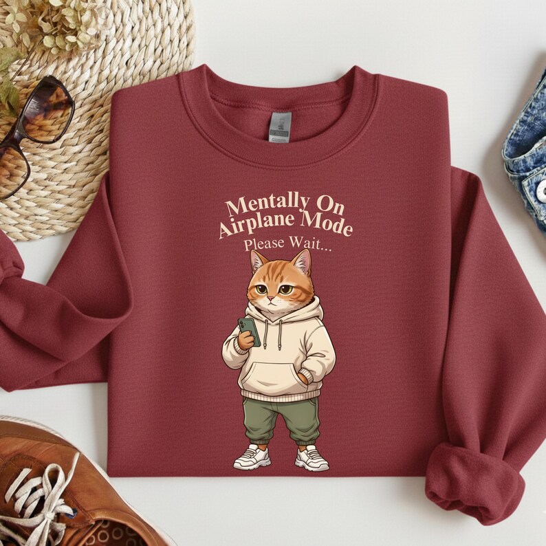 May include: A maroon sweatshirt with the text "Mentally On Airplane Mode Please Wait..." above a cartoon cat wearing a hoodie, green pants, and sneakers, holding a phone. The sweatshirt is folded.