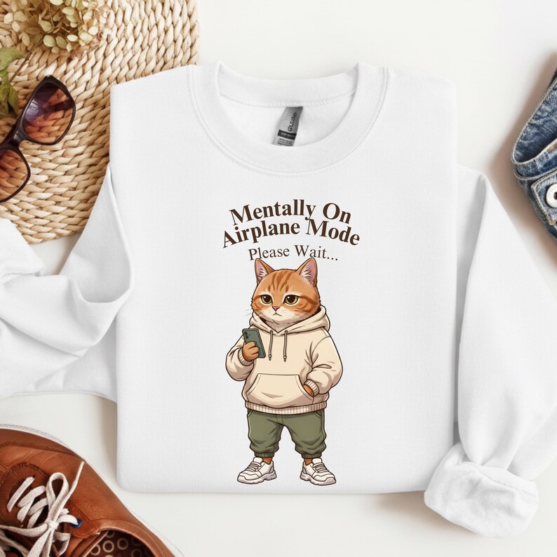 May include: White sweatshirt with the text "Mentally On Airplane Mode Please Wait..." and a cartoon cat wearing a hoodie, green pants, and sneakers, holding a phone.
