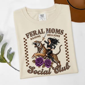 May include: Cream-colored t-shirt with the text "FERAL MOMS RAISING HELLIONS Social Club." The graphic features a woman riding a tiger, holding a sword, with purple roses at the bottom. The shirt has a brown checkered pattern on the sides.