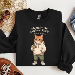 May include: Black sweatshirt featuring a cartoon cat wearing a hoodie and holding a phone. The cat is orange and white, with green pants and white sneakers. Text on the sweatshirt reads "Mentally On Airplane Mode Please Wait..."