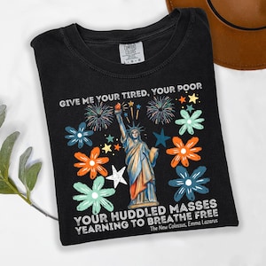 Puede incluir: Camiseta negra con un dise&ntilde;o de la Estatua de la Libertad, rodeada de flores y fuegos artificiales. El texto dice "GIVE ME YOUR TIRED, YOUR POOR" y "YOUR HUDDLED MASSES YEARNING TO BREATHE FREE".