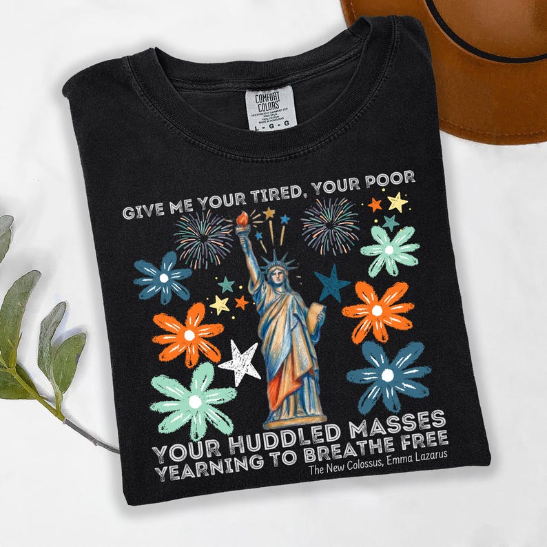 Puede incluir: Camiseta negra con un dise&ntilde;o de la Estatua de la Libertad, rodeada de flores y fuegos artificiales. El texto dice "GIVE ME YOUR TIRED, YOUR POOR" y "YOUR HUDDLED MASSES YEARNING TO BREATHE FREE".