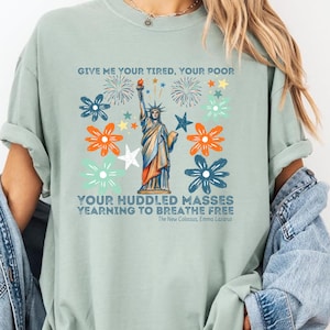 Puede incluir: Camiseta verde claro con un dise&ntilde;o de la Estatua de la Libertad, fuegos artificiales y elementos florales. El texto "GIVE ME YOUR TIRED, YOUR POOR" y "YOUR HUDDLED MASSES YEARNING TO BREATHE FREE" est&aacute; impreso.
