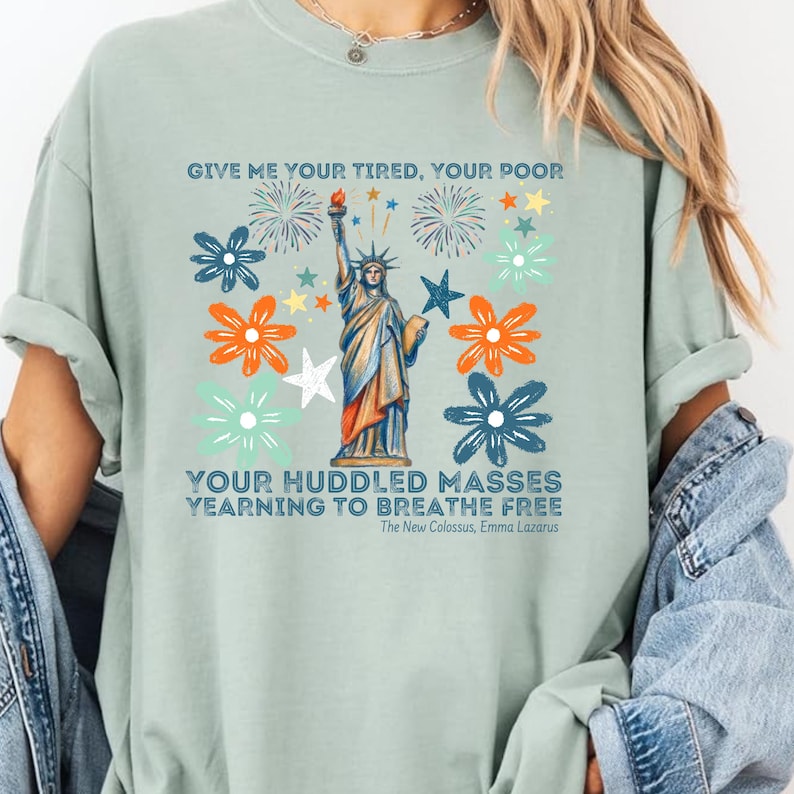 Puede incluir: Camiseta verde claro con un dise&ntilde;o de la Estatua de la Libertad, fuegos artificiales y elementos florales. El texto "GIVE ME YOUR TIRED, YOUR POOR" y "YOUR HUDDLED MASSES YEARNING TO BREATHE FREE" est&aacute; impreso.