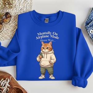 May include: A royal blue sweatshirt featuring a cartoon cat wearing a hoodie and holding a phone. The text "Mentally On Airplane Mode Please Wait..." is printed above the cat. The cat is wearing green pants and white sneakers.