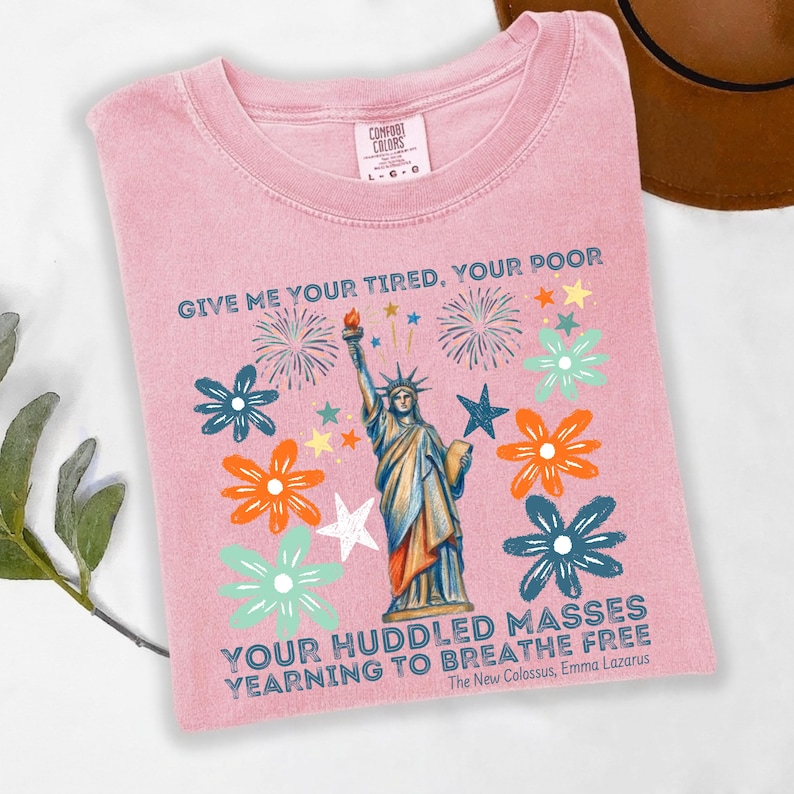 Puede incluir: Camiseta rosa Comfort Colors con un gr&aacute;fico de la Estatua de la Libertad, fuegos artificiales y flores. El texto dice: "Give me your tired, your poor... Yearning to breathe free." Un sombrero marr&oacute;n es visible en el fondo.
