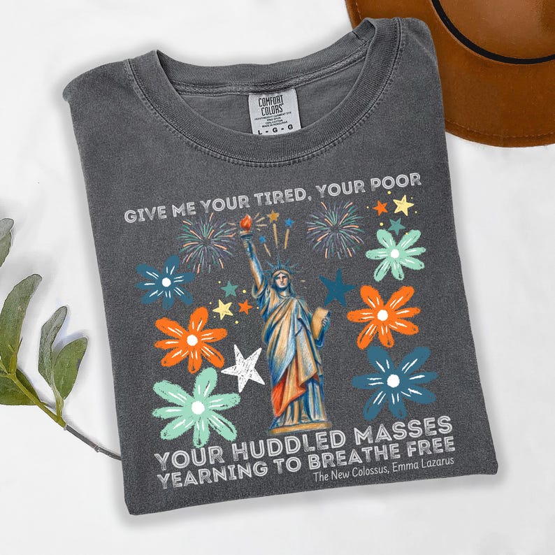 Puede incluir: Camiseta gris oscuro con un dise&ntilde;o de la Estatua de la Libertad, fuegos artificiales y flores. El texto en la camiseta dice: "GIVE ME YOUR TIRED, YOUR POOR, YOUR HUDDLED MASSES YEARNING TO BREATHE FREE" de The New Colossus de Emma Lazarus.