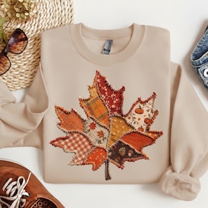 May include: A beige crewneck sweatshirt featuring a large, central maple leaf graphic composed of various fabric-style patches in shades of orange, red, brown, and cream, with patterns including gingham, floral, and pumpkin motifs.