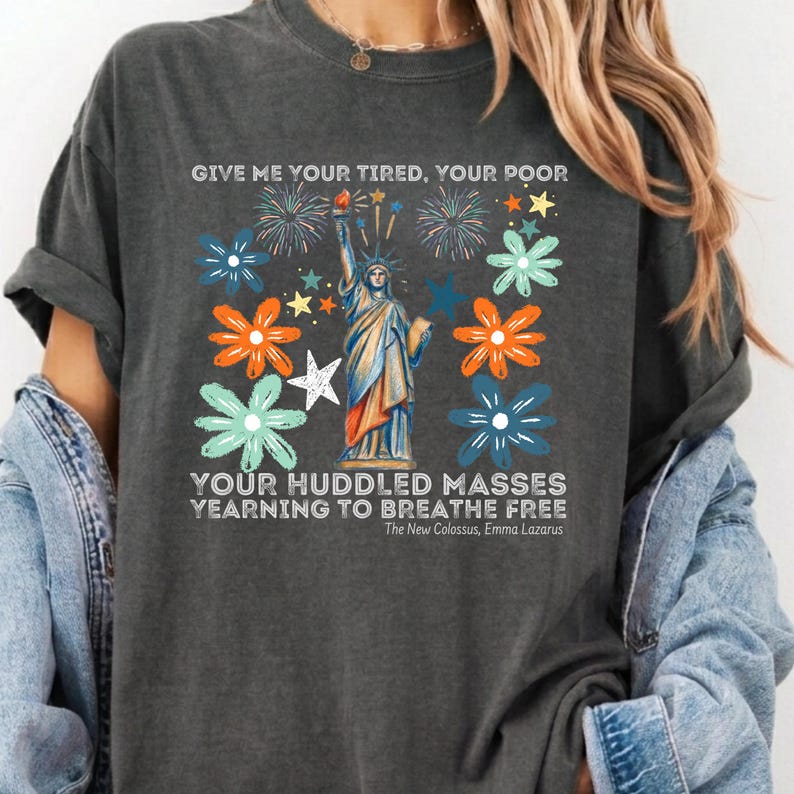 Puede incluir: Camiseta gris oscuro con un gr&aacute;fico de la Estatua de la Libertad, fuegos artificiales y detalles florales. El texto en la camiseta dice: "GIVE ME YOUR TIRED, YOUR POOR...YEARNING TO BREATHE FREE."