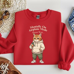 May include: Red sweatshirt with the text "Mentally On Airplane Mode Please Wait..." and a cartoon cat wearing a hoodie and holding a phone. The cat is wearing green pants and white sneakers.