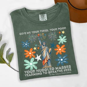 Puede incluir: Una camiseta verde Comfort Colors con un dise&ntilde;o de la Estatua de la Libertad, fuegos artificiales y dise&ntilde;os florales. La camiseta muestra el texto "GIVE ME YOUR TIRED, YOUR POOR" y "YOUR HUDDLED MASSES YEARNING TO BREATHE FREE."
